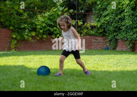 Das kleine Mädchen spielt mit ihrem blauen Ball im Garten ihres Hauses. Mädchen Mit Selektivem Fokus Stockfoto