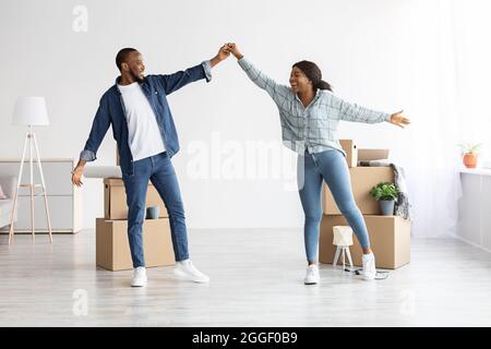 Beweglicher Tag. Fröhliche Afroamerikanische Ehepartner, Die In Ihrer Neuen Wohnung Zwischen Kartons Tanzen, Feiern Glückliche Junge Schwarze Ehepartner Den Umzug Nach Stockfoto