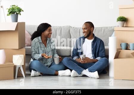 Glückliches Schwarzes Paar, Das Sich Unter Kartons Mit Habseligkeiten Entspannt Hat Nach Dem Umzug In Die Neue Wohnung, Fröhliche Afroamerikanische Kaffeetrinkerin Und Plauderin, Re Stockfoto