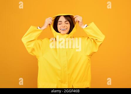 Aufgeregt Millennial Dame in gelber wasserdichter Regenjacke, die Kapuze aufsetzt, auf gelbem Hintergrund steht und mit geschlossenen Augen lächelt. Herbst kaltes Wetter Stockfoto