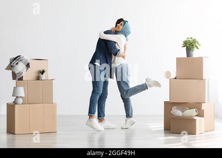 Emotionale junge muslimische Dame in Hijab umarmte ihren Mann, Mann und Frau aus dem Nahen Osten, die zwischen Papierkisten und anderen Dingen im leeren Raum, der Familie, stehen Stockfoto