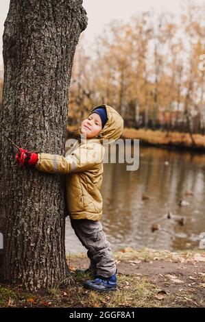 Der kleine glückliche, vierjährige Junge, der seine Augen zudrückte, umarmt in Herbstkleidung einen Baumstamm während eines Spaziergangs im Herbstpark in der Nähe des Teiches Stockfoto