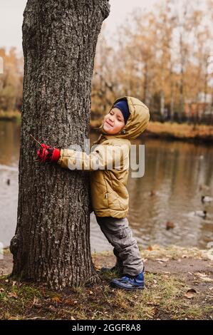 Der kleine glückliche, vierjährige Junge, der seine Augen zudrückte, umarmt in Herbstkleidung einen Baumstamm während eines Spaziergangs im Herbstpark in der Nähe des Teiches Stockfoto