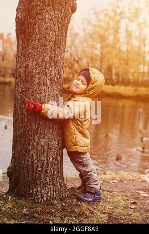 Kleiner glücklicher, vierjähriger Junge, der seine Augen zudrückte, umarmt in Herbstkleidung einen Baumstamm während eines Spaziergangs im Herbstpark in der Nähe des Teiches. Flar Stockfoto