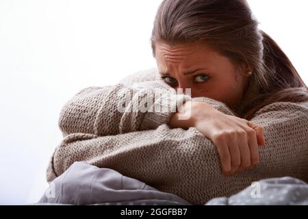 Unruhige oder kranke junge Frau, die ihr Gesicht in ihrem Arm versteckt und die Kamera mit weiten Augen und einem ängstlichen Stirnrunzeln in einem abgeschnittenen Nahaufnahme-Porträt anschaut Stockfoto