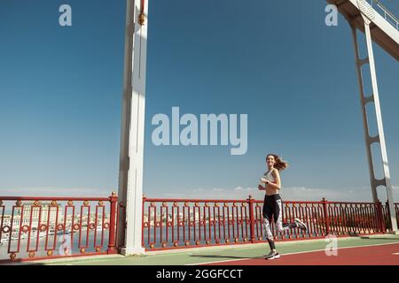 Lady mit fliegenden Haaren waering stilvoller Trainingsanzug läuft entlang des modernen Steges Stockfoto