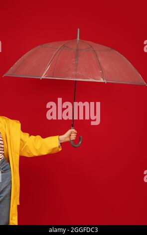 Frau im Regenmantel, die den transparenten Regenschirm gegen den roten Hintergrund, die Aufnahme im Studio und die Ernte hielt. Weibliche Hand hält stilvolle Sonnenschirm. Herbstzubehör Stockfoto