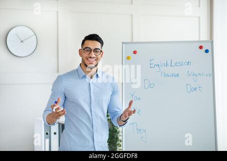 Lächelnder arabischer Lehrer, der fernab Unterricht gibt, in der Nähe einer Tafel steht und den Schülern vom Heimbüro die englischen Regeln erklärt. Online-Nachhilfe und dist Stockfoto