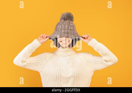 Überfreuliche Dame, die grauen Wollhut herunterzieht und lächelt, täuscht und vor der Kamera auf gelbem Hintergrund posiert. Glückliche Dame im heißen Winter Outfit wird s Stockfoto