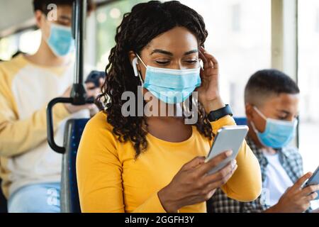 Konzept Der Online-Kommunikation. Porträt einer glücklichen jungen afroamerikanischen Frau in chirurgischer Maske Hören von Musik in drahtlosen Ohrhörern sitzen in Bus, L Stockfoto