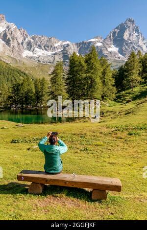 Eine Frau, die auf einer Holzbank sitzt, fotografiert den Blauen See und den Matterhorn-Berg, die sich auf dem Wasser, im Aostatal, Italien, spiegeln Stockfoto