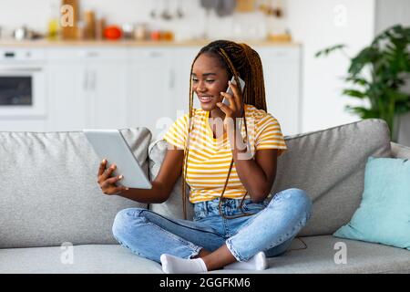 Fröhliche afroamerikanische Freiberuflerin, die auf dem Mobiltelefon spricht und auf ein digitales Tablet schaut, von zu Hause aus während der Sperre arbeitet, Speicherplatz kopieren. Glücklich af Stockfoto