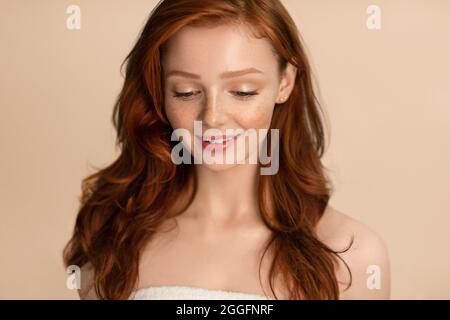 Attraktive Redhead Junge Frau Stehend In Handtuch Nach Der Dusche Gewickelt Posiert Blick Nach Unten Über Beige Studio Hintergrund. Natürliche Weibliche Schönheit Und Körper Stockfoto