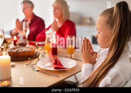 Kleines Mädchen, das am Festtisch sitzt, vor dem Familienessen mit ihren Großeltern betet, Gott dankt, den Urlaub mit ihren Ohren feiert Stockfoto