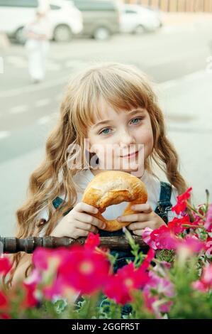 Schönes blauäugiges Mädchen mit einer Brezel auf der Straße. Porträt eines blauäugigen Mädchens mit langen goldenen Haaren. Stockfoto