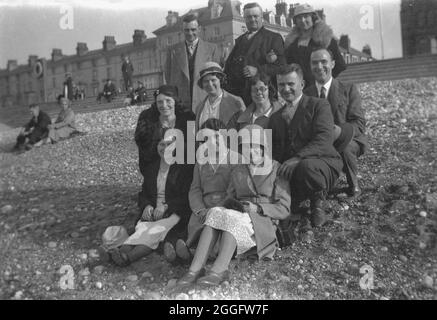 1930, historisch, versammeln sich zwei Familien an einem steinigen Strand für ein Gruppenfoto. Sie tragen die formelle Kleidung der Zeit, mit den Männern in Anzügen und Krawatten und den Damen in Kleidern und Hüten, England, Großbritannien. Stockfoto