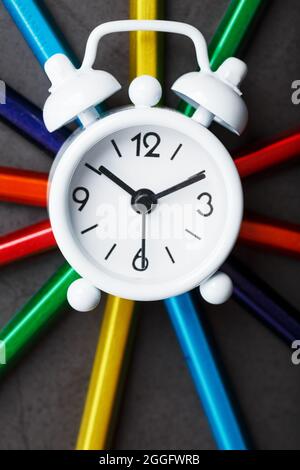Weißer Wecker auf Bleistiften in verschiedenen Farben in Sternform auf schwarzem Hintergrund. Das Konzept von Kreativität und Studienzeit Stockfoto