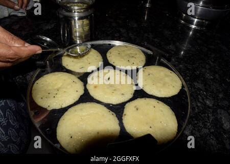 Weibliche Küche machen beliebte südindische gedünstete Gericht rava idli oder Grieß idli. Ghee oder klarierte Butter auf idlis gießen.Mutter kocht vorbereiten Stockfoto