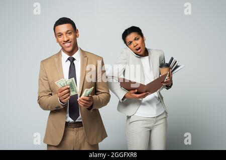 Glücklicher afroamerikanischer Geschäftsmann, der Dollar in der Nähe zählt, schockierte Sekretärin, die auf dem Smartphone auf Grau sprach Stockfoto