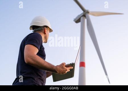Ein Ingenieur im Helm steuert und wartet den Betrieb einer Windenergieanlage mit einem Laptop auf dem Hintergrund der Windmühle Stockfoto