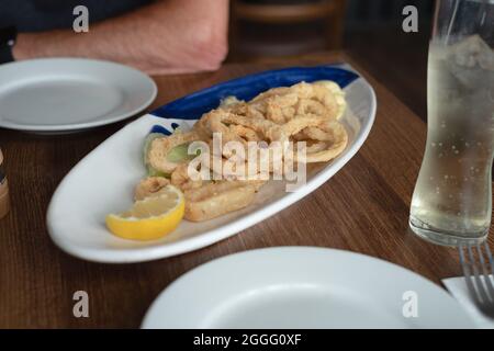 Calamari-Ringe auf einem ovalen Teller auf einem Tisch in einem Restaurant. Das Gericht wird zum Mitteilen serviert. Es gibt zwei kleine Teller für die Gäste. Arm eines Mannes Stockfoto