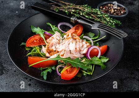 Salat mit gebratenem Lachsfiletsteak, frischem Salatrugula und Tomate auf einem Teller. Schwarzer Hintergrund. Draufsicht Stockfoto