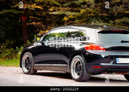 Rückansicht Foto eines luxuriösen schwarzen glänzenden Automobils mit coolen Rädern, das im Wald auf der Straße steht und keinen Fahrer im Auto hat. Stockfoto