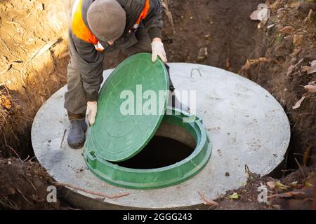 Ein Arbeiter installiert einen Kanalschacht an einem Klärbecken aus Betonringen. Bau von Kanalisationsnetzen für Landhäuser. Stockfoto