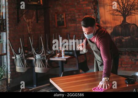 Der junge Kellner desinfiziert den Tisch im Café, während er Gesichtsmaske trägt Stockfoto