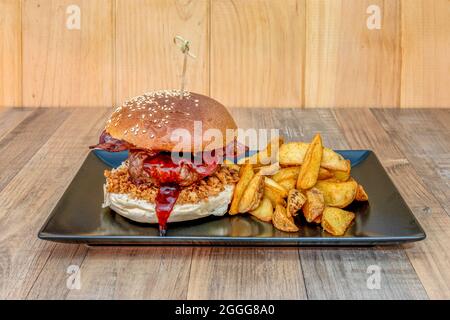 Rindfleischburger mit Speck, auf knuspriger Zwiebel, beträufelter Erdbeermarmelade und mit gebratenen Kartoffelkeilen mit Garnierung auf der Haut Stockfoto