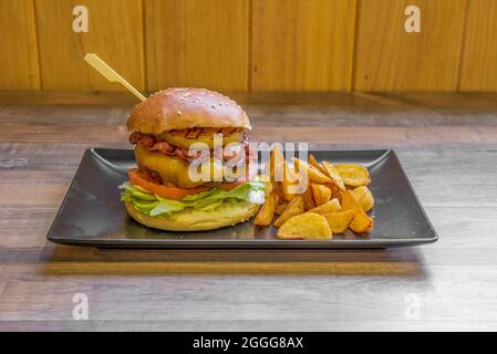 Rinderburger mit Salatblättern, eingelegten Gurken, gebratenem Speck in Scheiben, Spiegelei mit dem Eigelb und Kartoffelkeil auf einem schwarzen Platt garnieren Stockfoto