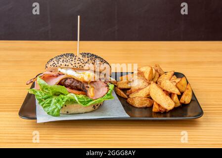 Rinderburger mit Salatblättern, eingelegten Gurken, gebratenem Speck in Scheiben, Spiegelei mit dem Eigelb und Kartoffelkeil auf einem schwarzen Platt garnieren Stockfoto