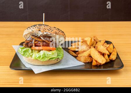 Klassischer Mayonnaise-Burger mit Salat, Rindfleisch, karamellisierten Zwiebeln und Tomatenscheiben mit Mohnsamen, garniert mit Kartoffelkeilen Stockfoto