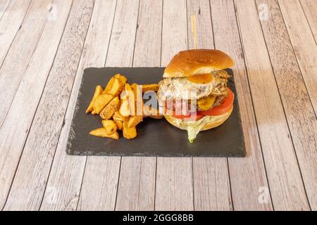 Klassischer Rindfleischburger mit Spiegelei, gebratenem Speck in Scheiben, geschmolzenem Cheddar-Käse, Tomatenscheiben und Gurken sowie Salatblättern, garniert mit gebratenem Potat Stockfoto