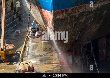 Arbeiter, die die Korrosion des Rumpfes eines Handelsschiffs mit Drucksandstrahlen in einem Trockendock einer modernen Werft reinigen Stockfoto