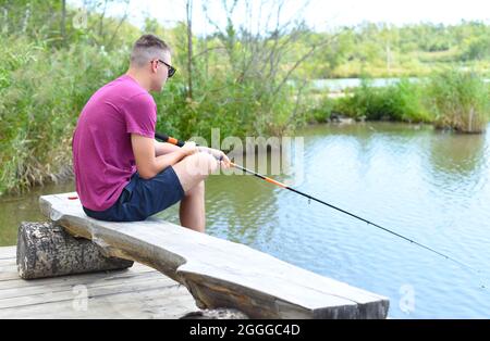 Junger Fischer sitzt auf hölzernen Pier, Angeln im See. Er hält die Rute und schaut auf Schwimmer Stockfoto