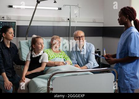 afroamerikanische Krankenschwester diskutiert Krankheitssymptome mit älteren kranken älteren Frauen auf der Krankenstation. Fröhliche Familie besucht eine pensionierte Patientin, die sie während der klinischen Genesung unterstützt. Medizinische Therapie Stockfoto