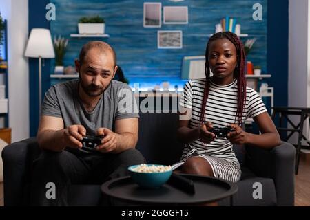 POV von interracial paar spielen Videospiel auf Konsole mit Joysticks im Fernsehen. Multiethnische Partner blicken auf die Kamera und genießen dabei lustige Aktivitäten mit Technologie und Controllern Stockfoto