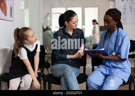 Familie diskutiert medizinische Behandlung mit afroamerikanischen Krankenschwester im Krankenhaus Warteraum während klinischer Termin. afroamerikanische Krankenschwester schreibt klinische Expertise. Support-Service im Gesundheitswesen Stockfoto
