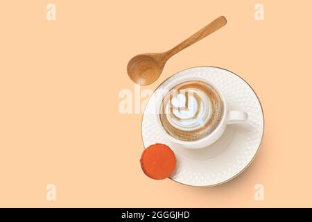 Eine weiße Porzellanschale mit Cappuccino-Kaffee und eine goldgerahmte Untertasse mit einem Holzlöffel und einem orangefarbenen Makronen schwebendem. Stockfoto