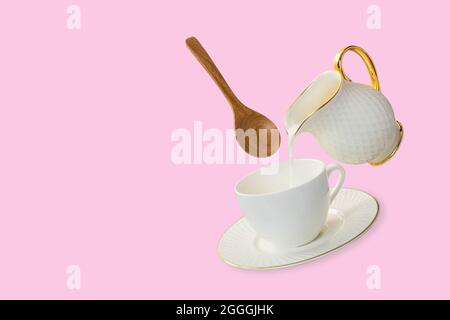 Levitation weiße Kaffeetasse und Untertasse mit Milch, die aus einem Milchkännchen mit einem schwimmenden Holzlöffel ausgegossen wird. Isolation auf einem weichen rosa Hintergrund. Levita Stockfoto