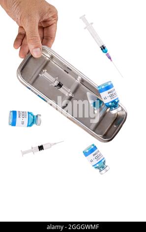 Eine Hand, die eine Metallschale mit fallenden Glasfläschchen mit der Bezeichnung Covid-19-Impfstoff und mehreren Spritzen hält. Isolation auf weißem Hintergrund. Stockfoto
