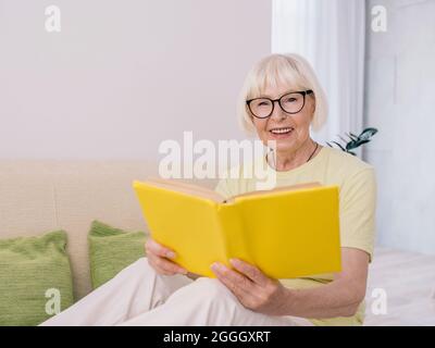Ältere Frau mit grauen Haaren liest zu Hause ein Buch von einem Sofa. Bildung, Rente, Anti-Age, Lesekonzept Stockfoto