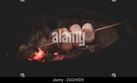 Drei Marschmalchen auf einem Stock werden gegrillt. Glühende Kohlen sind darunter zu sehen. Stockfoto