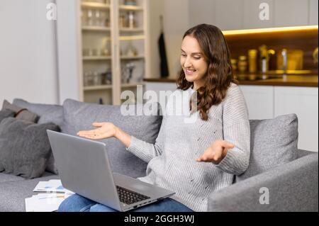 Eine fröhliche junge Frau sitzt zu Hause auf der Couch und unterhält sich online mit einem Laptop für Online-Gespräche. Videoanruf-Konzept Stockfoto