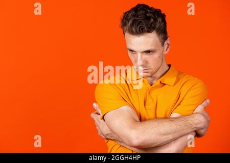 Junger Mann, der mit gekreuzten Händen auf orangefarbenem Studiohintergrund steht. Konzept der menschlichen Emotionen, Mimik, Jugend, Gefühle, Werbung. Stockfoto