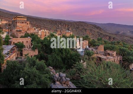 Altes Bergdorf Misfat im Oman der Arabischen Halbinsel bei Sonnenuntergang. Alte Schlammhütten des Dorfes in einer Oase mit Palmen auf einem Berghang. Stockfoto