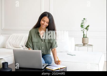 Indische Studentin, die online lernt, sich ein Webinar am Laptop anschaut, sich über das Thema aufhält und sich Notizen macht zu Hause auf der Couch sitzend, blickt die östliche Frau auf den Bildschirm und schreibt in das Notizbuch Stockfoto
