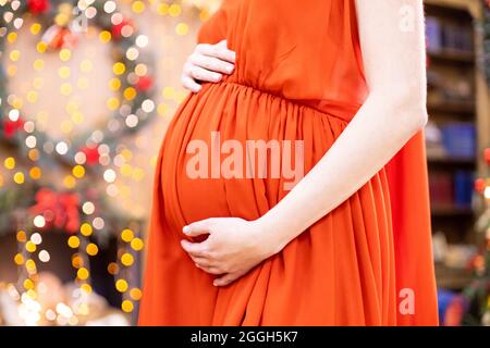 Bestes weihnachtsgeschenk. Gesichtslose Schwangere in einem roten Kleid umarmt ihren Bauch vor dem Hintergrund des Weihnachtswohnzimmers in Unschärfe. Nahaufnahme kein fa Stockfoto