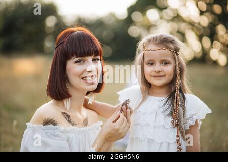 Nahaufnahme einer ziemlich lächelnden jungen Mutter und ihrer niedlichen Tochter im Boho-Stil mit Federn im Haar, die im Sommerfeld vor der Kamera posiert. Mama hält einen kleinen wilden Frosch in den Händen Stockfoto